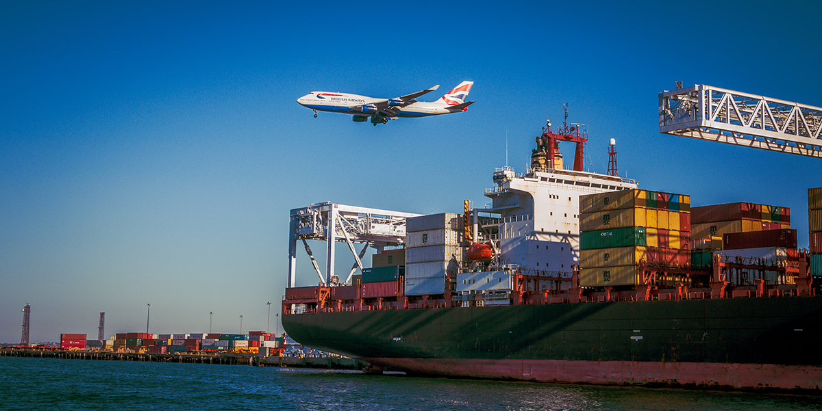 Aeroplane flying over container ship. Photo by VanveenJF on Unsplash